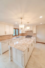 A wide angle photo from the right side of a contemporary kitchen remodel with a white and sand color scheme, two chandeliers hung over the kitchen island, and natural stone countertops