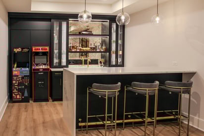 Wide angle photo of a home bar in the basement with three pendant lights suspended above and a decorative bar shelf mounted on the wall