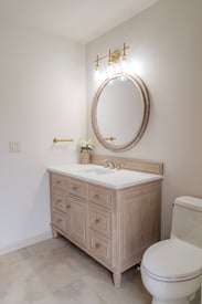 Single vanity powder room with white walls, brushed gold finished, a large round mirror, and the toilet on the right side