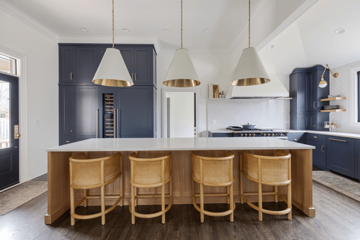 A natural wood center island with white quartz countertops, three conical pendant lights in white and gold contrast a kitchens navy blue cabinetry