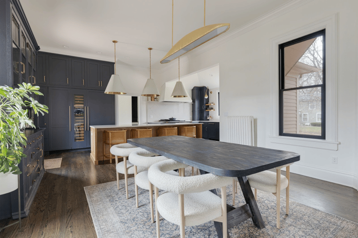 An open-plan kitchen and dining space with navy blue cabinets, a light wood center island, white quartz countertops and walls with gold and bronze accents.