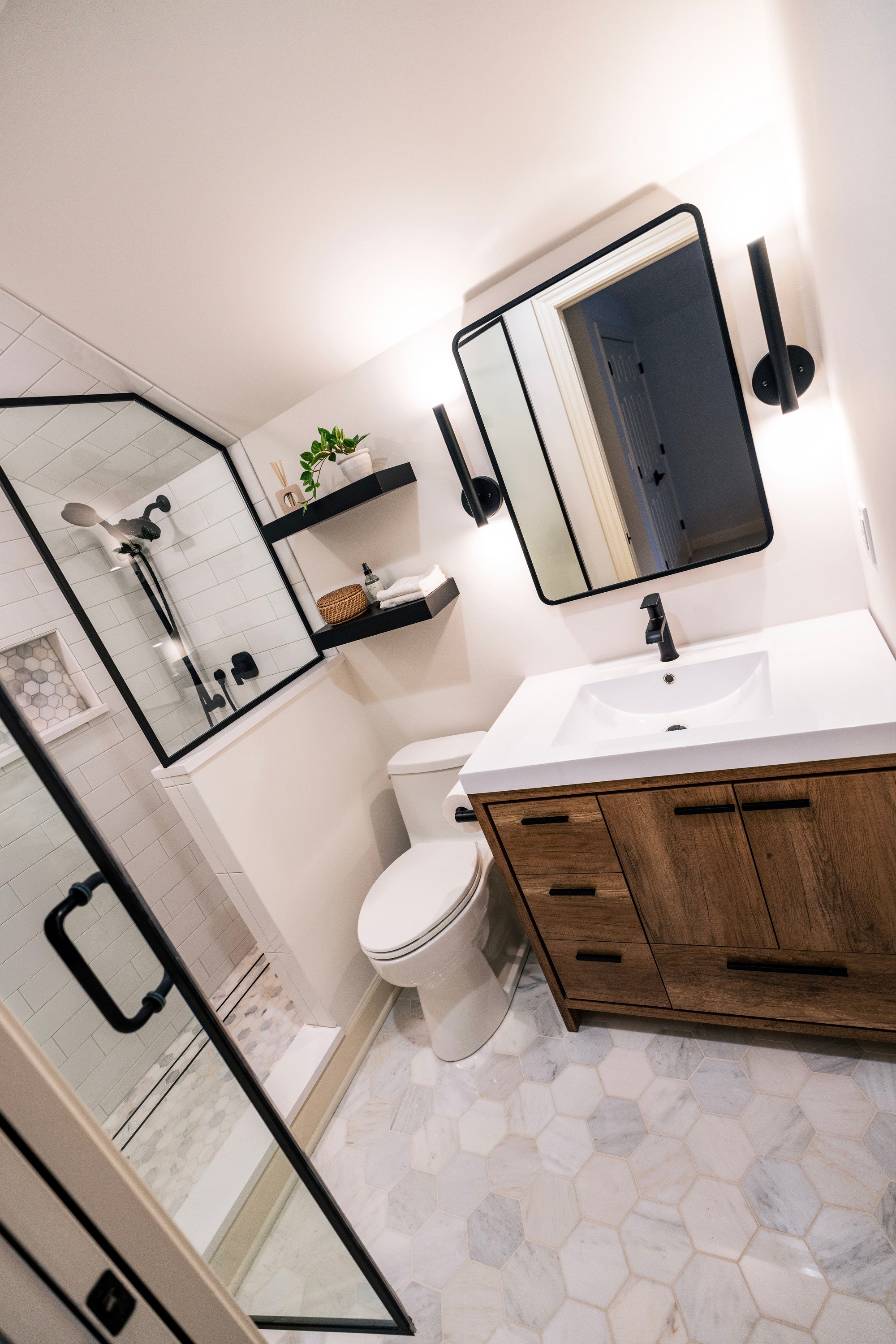 Elevated wide angle photo of the bathroom taken from the right hand corner of the room that shows the shower with the door open, the toilet, and single vanity all illuminated by LED lights